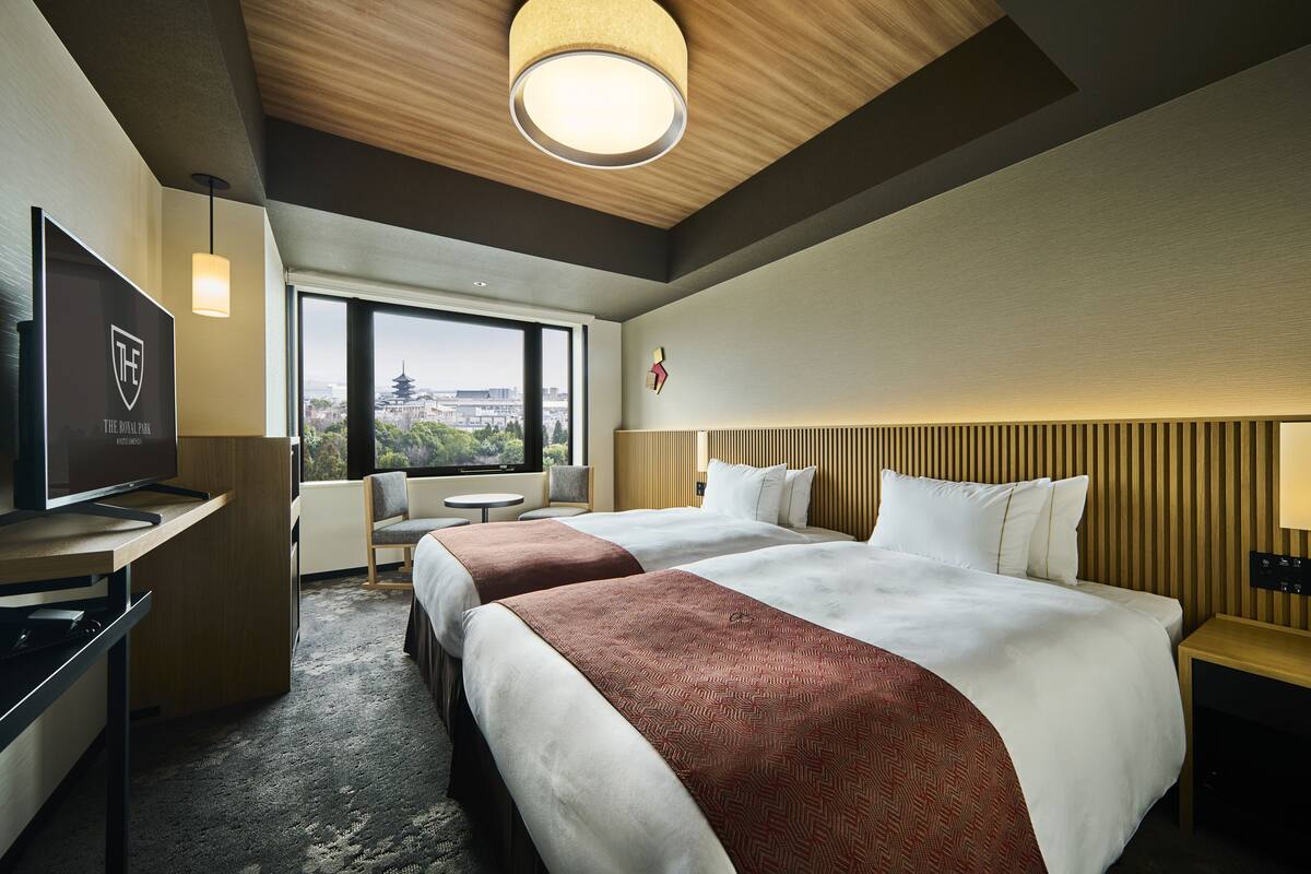 A hotel room with two beds, a desk with a TV, a chair, a view of the city, and a wooden ceiling.