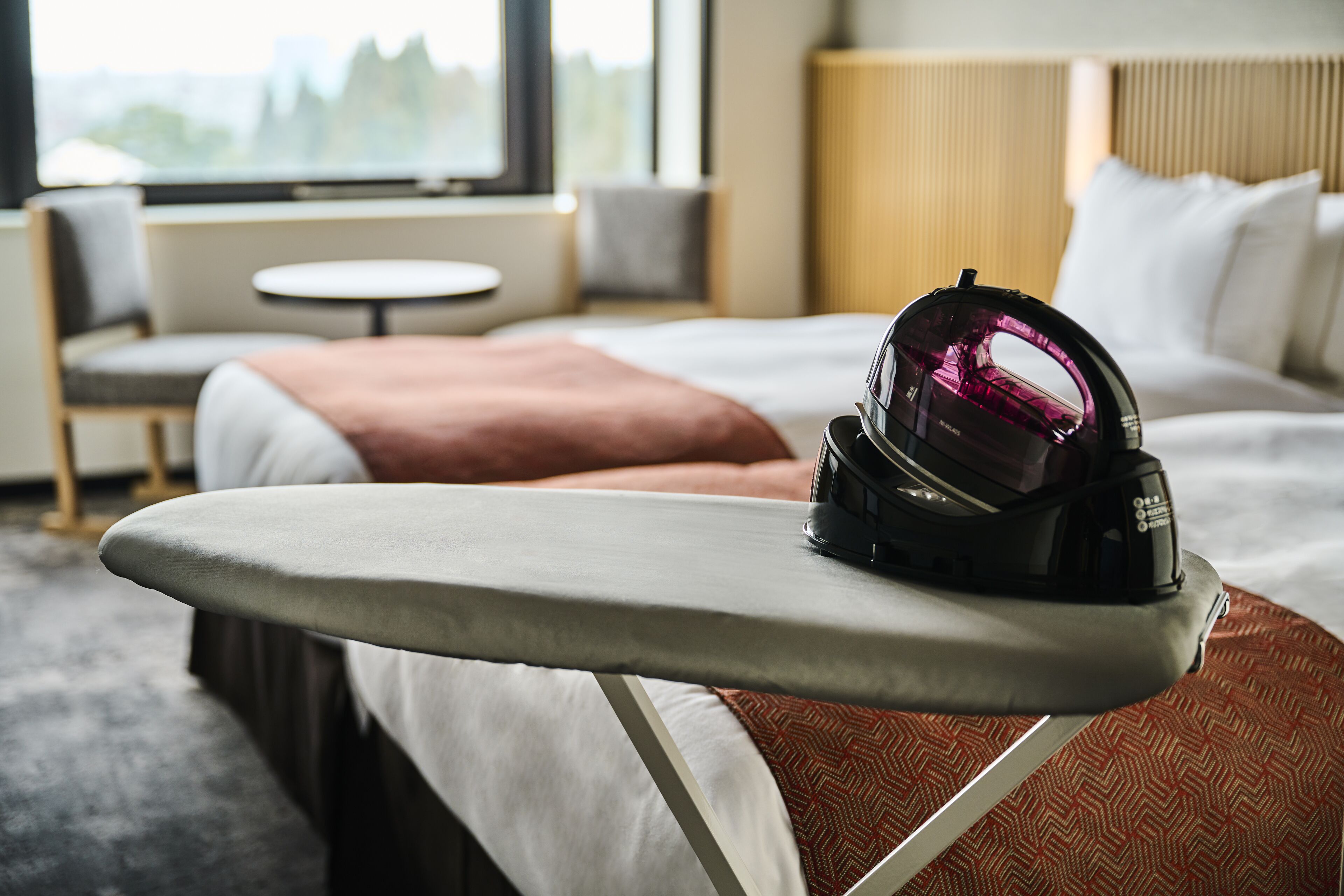 A neatly made bed with a pillow, a bedside table with an iron, and a window with a view.