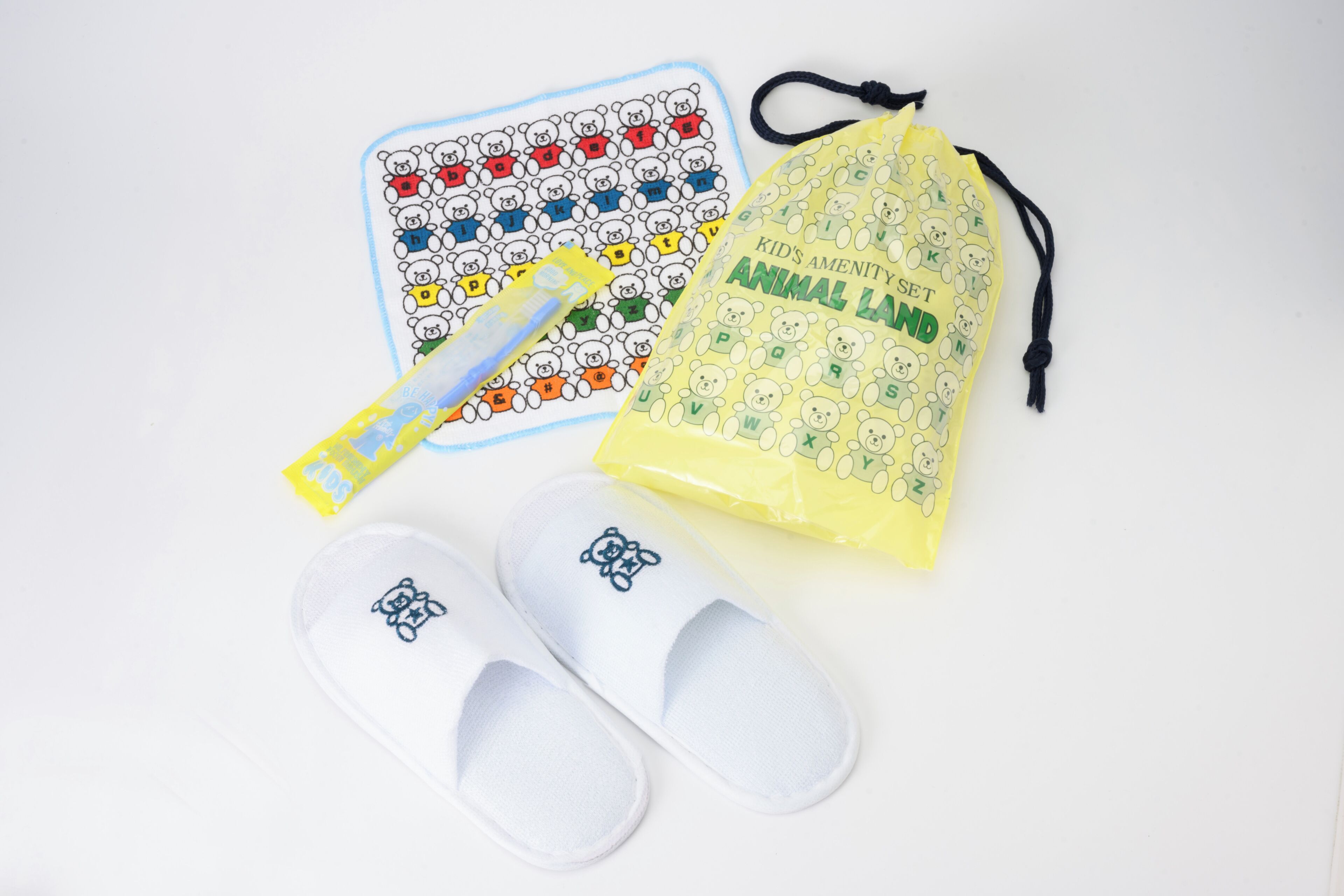 A pair of white slippers with a bear design, a yellow drawstring bag with bear patterns, and a blue and white alphabet-themed mat.