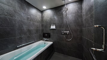Casa de banho moderna com azulejos escuros, uma banheira branca, um chuveiro manual e um painel de controlo fixado à parede.