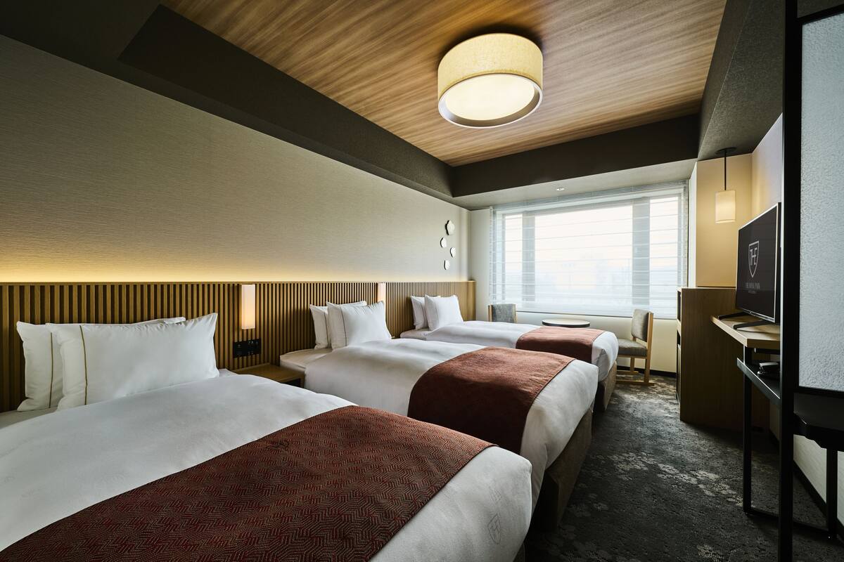 A hotel room with three beds, a desk, a TV, and a large window.