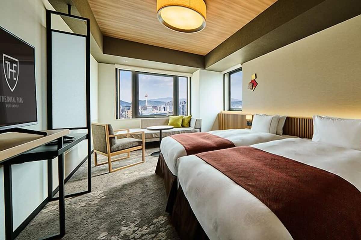 A hotel room with two beds, a desk, a chair, a small table, and a view of the cityscape.