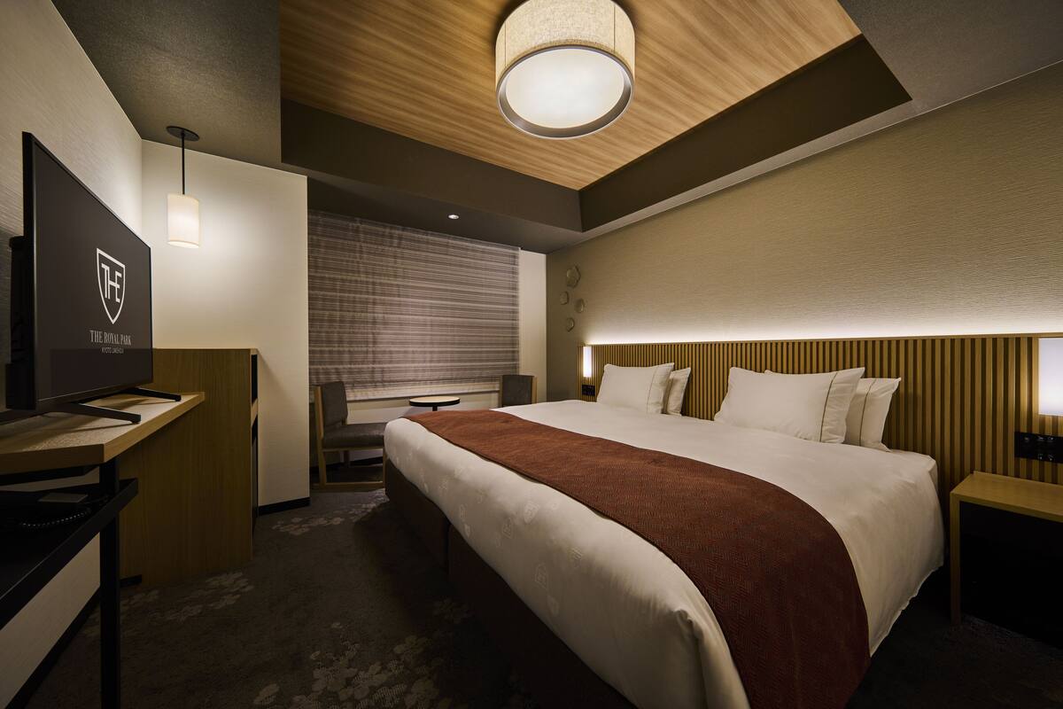 A hotel room with a large bed, a desk with a television, a lamp, and a window with blinds.