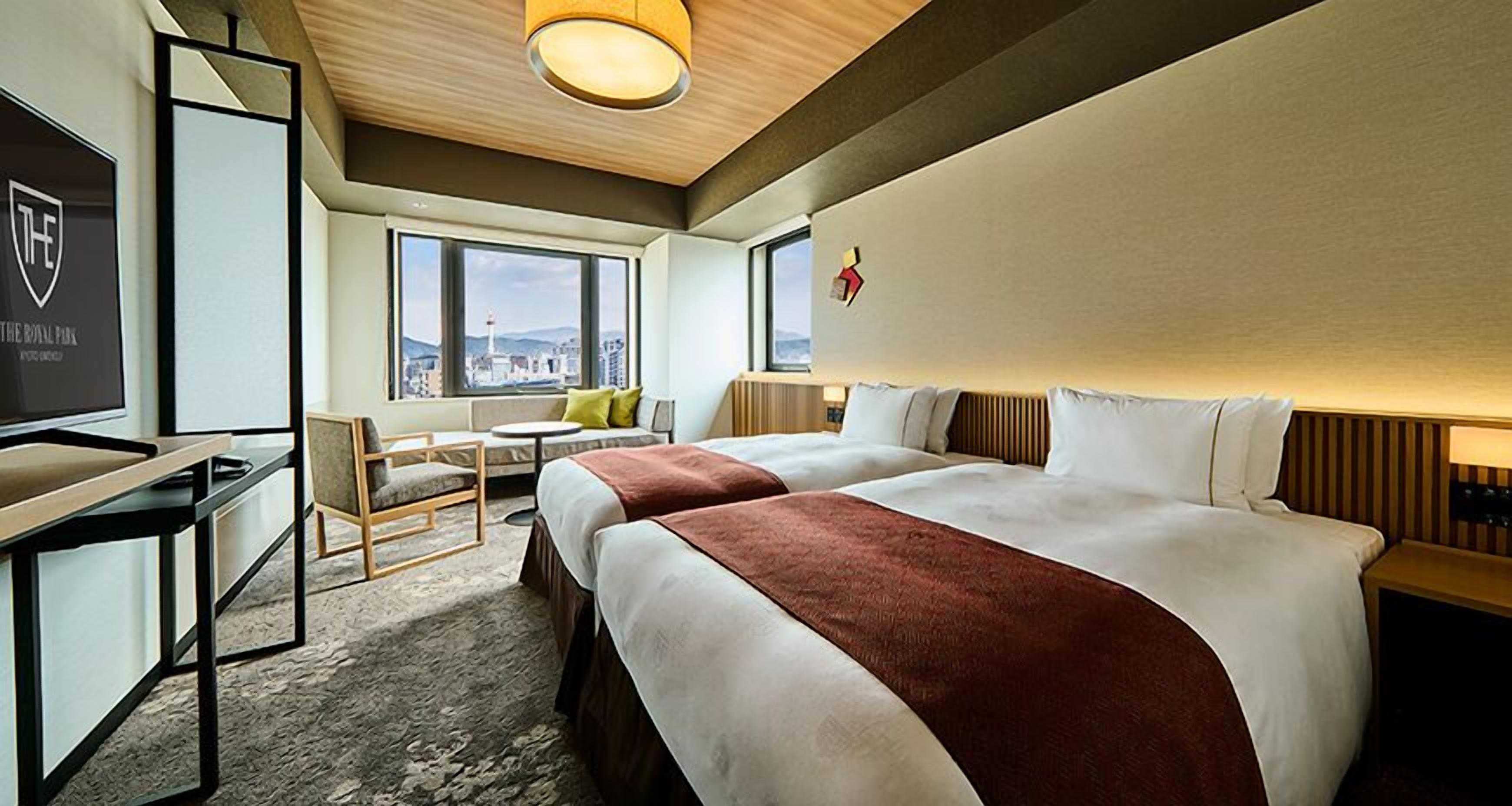 A hotel room with two beds, a desk, a chair, a small table, and a view of the city.