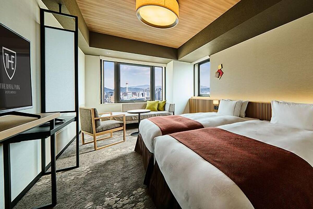 A hotel room with two beds, a desk, a chair, a small table, and a view of the city.