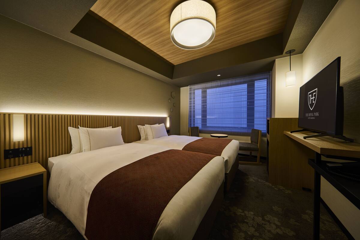 A hotel room with two beds, a desk, a television, and a window with blinds.