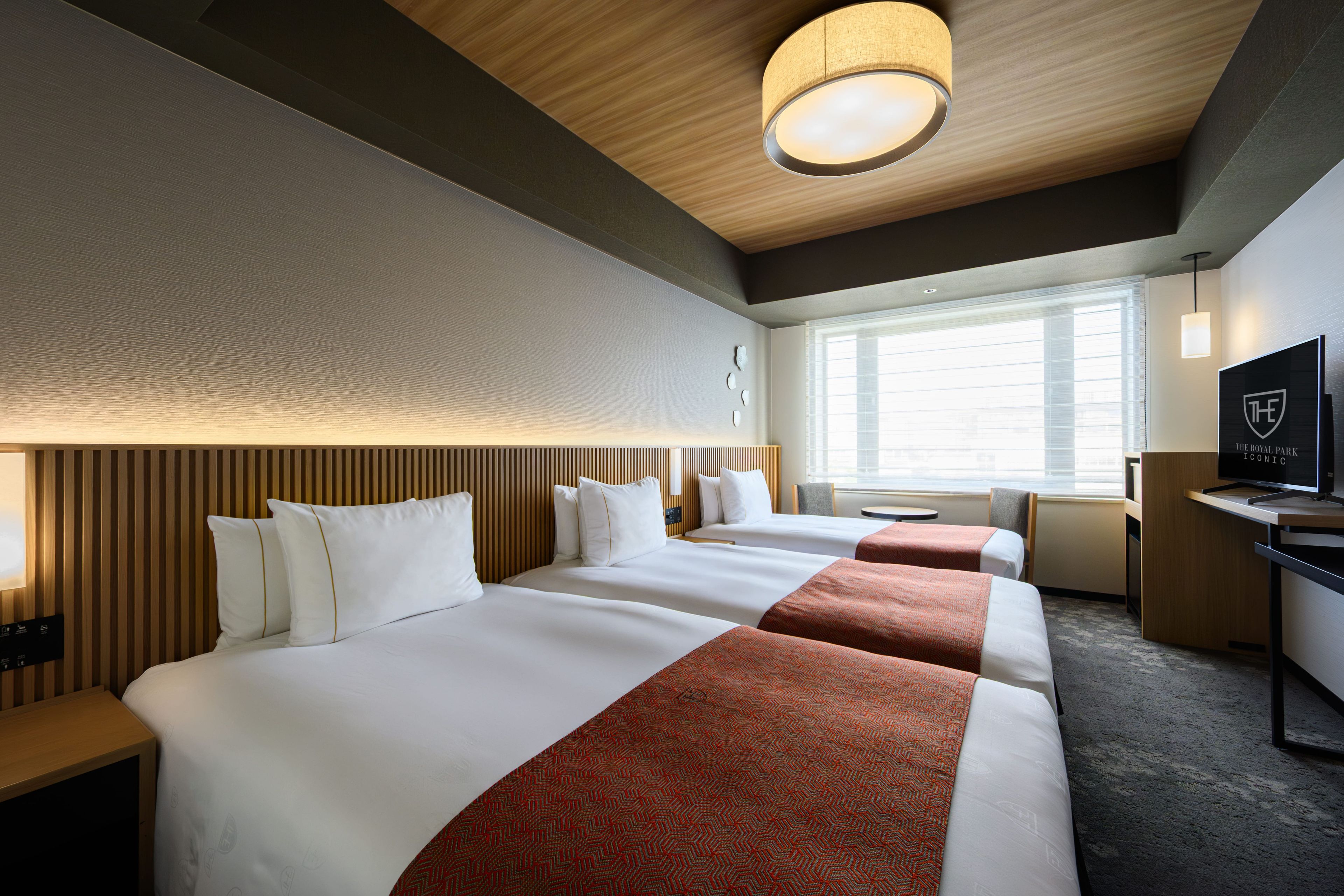 A hotel room with two beds, a desk, a television, and a window with blinds.