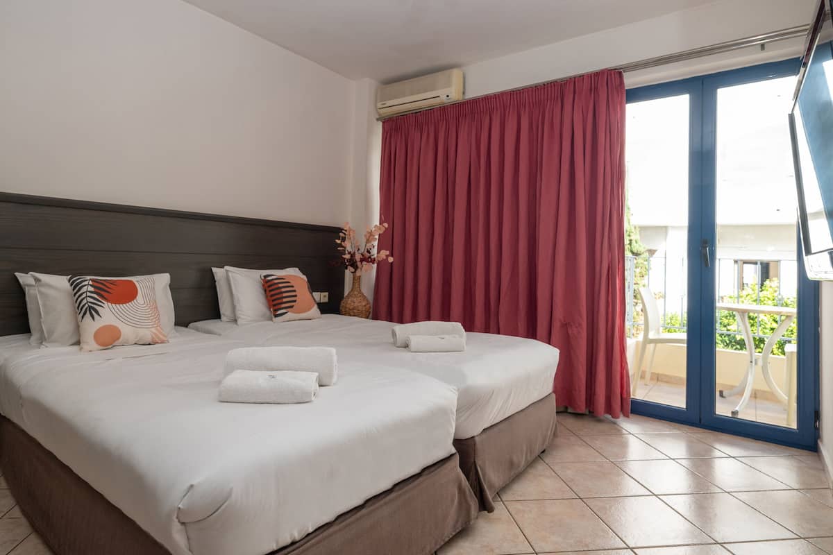 Cama doble con ropa blanca y almohadas decorativas, un ventanal con cortinas rojas y vistas al exterior.