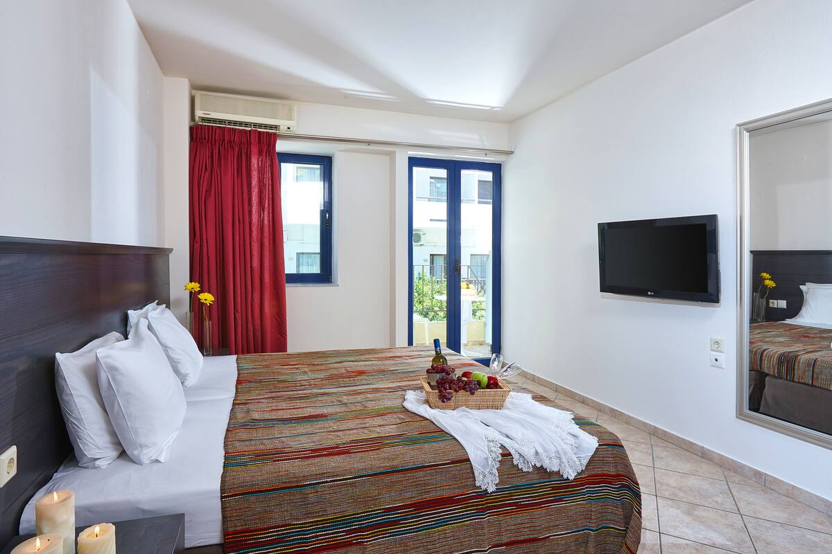 Habitación de hotel con cama, televisor, ventana con cortinas rojas y vistas al balcón.
