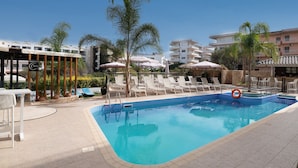 Una piscina con sillas de descanso y sombrillas, rodeada de palmeras y edificios.