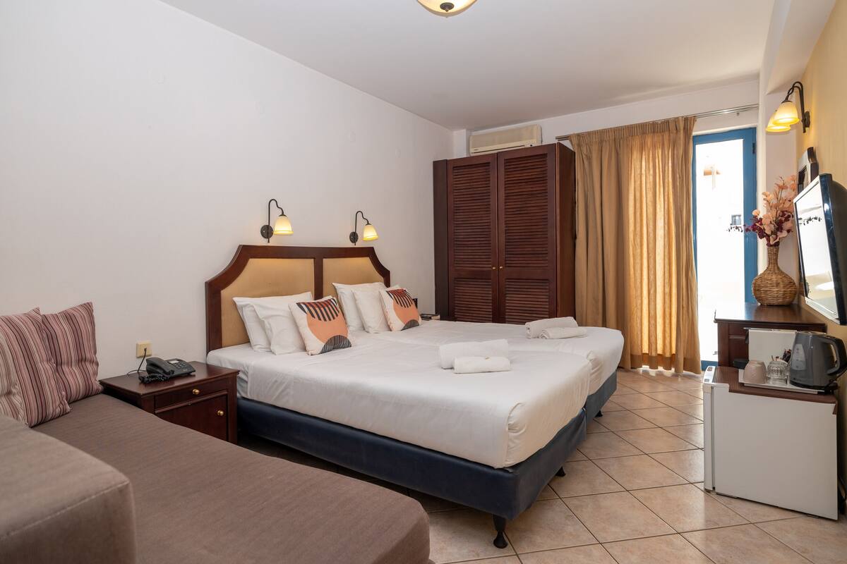 Habitación de hotel con una cama doble, una mesita de noche con lámpara, un armario y un pequeño escritorio con espejo y jarra.