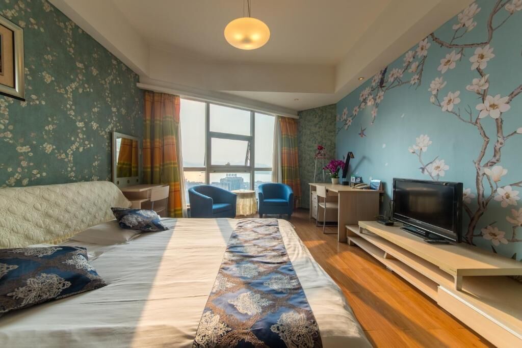 A hotel room with a large bed, a desk with a TV, and a view of the city through large windows.