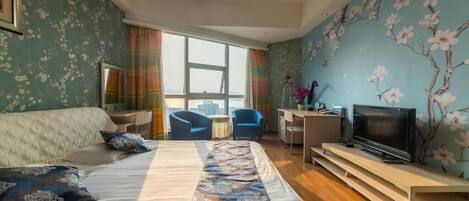 A hotel room with a large bed, a desk with a TV, and a view of the city through large windows.