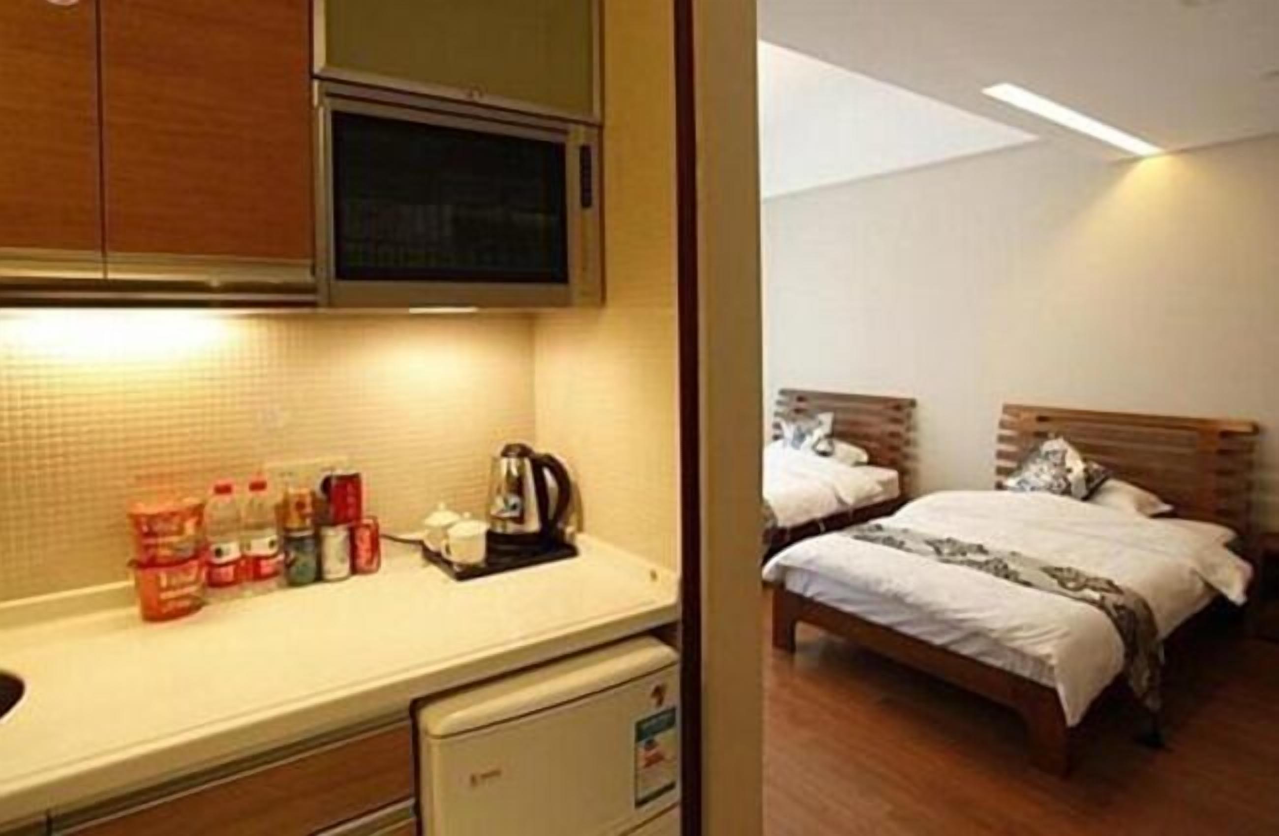 A compact hotel room with a kitchenette, a bed with two pillows, and a wooden headboard.