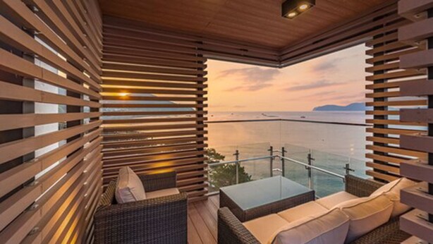 Ein Balkon mit Holzlamellen, Korbmöbeln und Blick auf das Meer bei Sonnenuntergang.