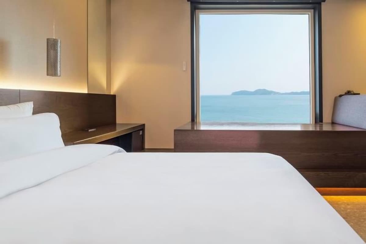 A hotel room with a large bed, a nightstand, a desk, and a view of the sea.