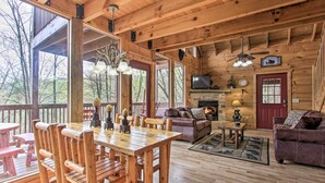 A rustic cabin interior with a wooden dining table, sofas, a fireplace, and large windows.