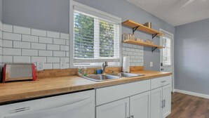 Een keuken met witte kastjes, een houten aanrecht, een spoelbak en een raam met jaloezieën.