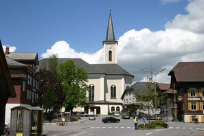 A church with a tall steeple and a clock, surrounded by buildings and a clear sky. - Hotel Engel (Alberschwende)