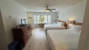 Un dormitorio con dos camas, un escritorio con computadora, un ventilador de techo y una ventana con vista a árboles.