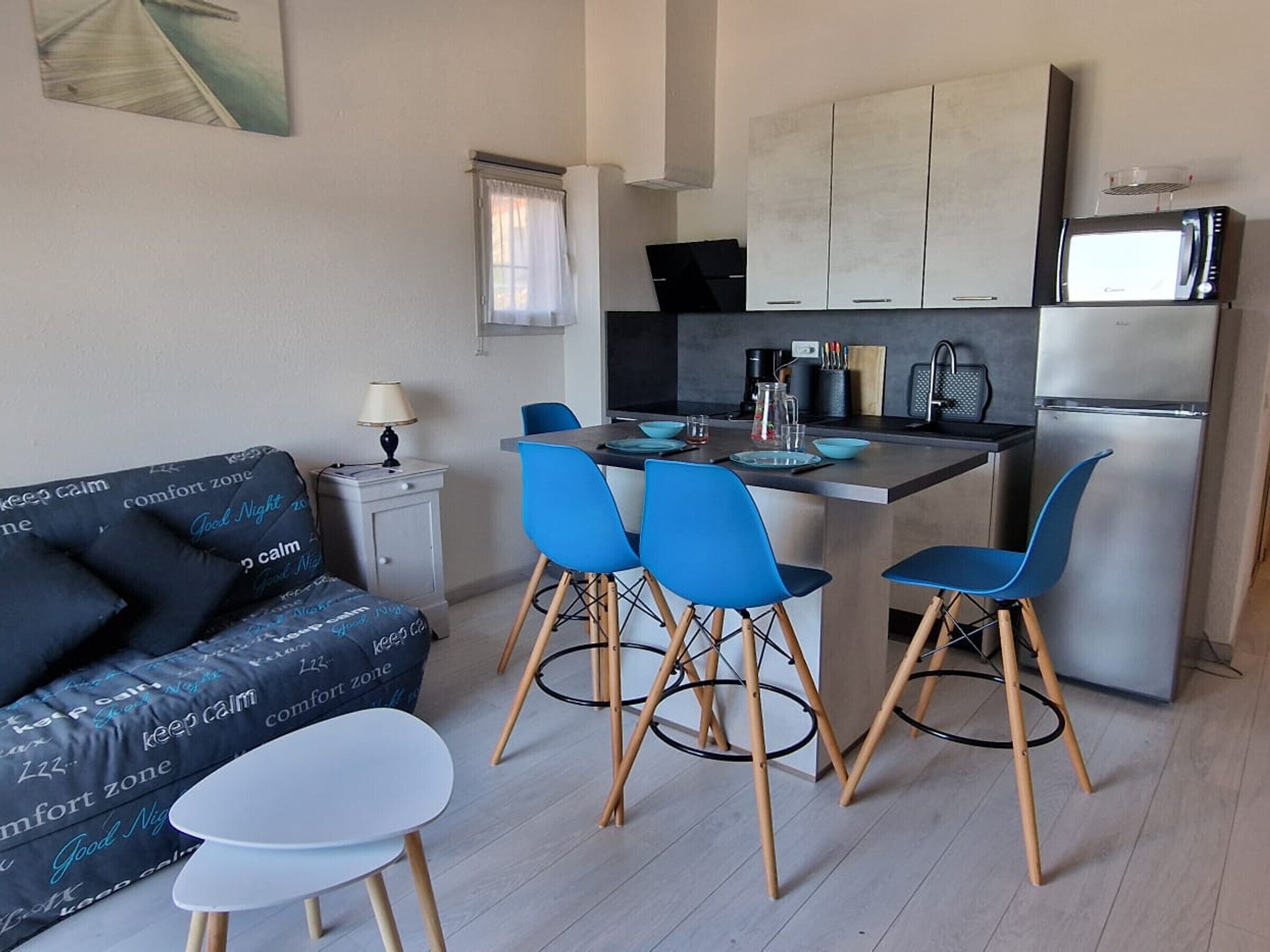 Un espacio habitable compacto con kitchenette, un sofá con cojines estampados, una pequeña mesa de comedor y sillas azules.
