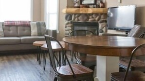 A living room with a stone fireplace, a sofa, and a dining table with chairs. - Spacious Townhouse / Loft Access / Mountain Views (Fernie)