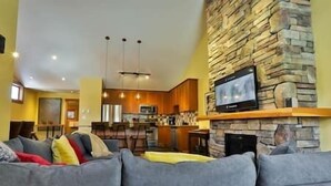 A living room with a grey sofa, a stone fireplace, and a kitchen in the background. - Alpine Chalet / Hot Tub / Garage (Fernie)