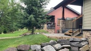 A modern house with a stone foundation, a large tree in the yard, and a well-maintained lawn. - Alpine Chalet / Hot Tub / Garage (Fernie)