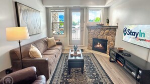 A living room with a sofa, coffee table, fireplace, TV, and a view of the outdoors through large windows. - 2 BDRM / Hot Tub & Pool / AC / Fireplace (Fernie)