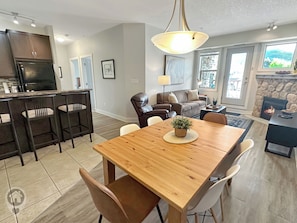 A modern kitchen with dark cabinets, a dining area with a wooden table and chairs, a leather sofa, and a stone fireplace. - 2 BDRM / Hot Tub & Pool / AC / Fireplace (Fernie)