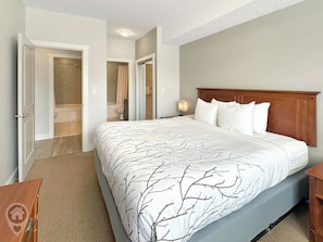 A bedroom with a large bed, a wooden headboard, a nightstand, and a bathroom visible through an open door. - Modern Fernie Apartment with Pool & Hot Tub (Fernie)