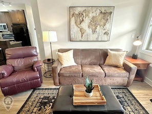 A living room with a leather sofa, a red chair, a coffee table with a plant, a world map on the wall, and a lamp. - 2 BDRM / Hot Tub & Pool / AC / Fireplace (Fernie)