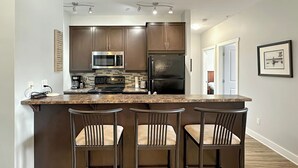 A kitchen with dark wooden cabinets, a stainless steel microwave, a black refrigerator, and a granite countertop. There are three bar stools with cushioned seats. - 2 BDRM / Hot Tub & Pool / AC / Fireplace (Fernie)
