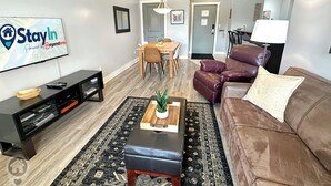 A living room with a TV, leather sofa, ottoman, dining table, chairs, and a rug. - 2 BDRM / Hot Tub & Pool / AC / Fireplace (Fernie)