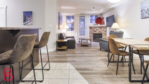 A modern living room with a fireplace, a dining area with a wooden table, and a kitchen area with a countertop. - Hot Tub & Sauna / 24-Hour Check-In / Quiet (Fernie)