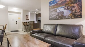 Ein modernes Wohnzimmer mit Ledersofa, einem Holztisch und einem Essbereich mit kleinem Tisch und Stühlen. Im Hintergrund ist eine Küche mit Mikrowelle und einer Bar erkennbar.