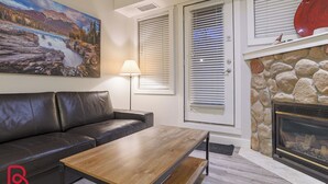 A living room with a black leather sofa, a wooden coffee table, a fireplace with a stone surround, and a painting of a waterfall. - Hot Tub & Sauna / 24-Hour Check-In / Quiet (Fernie)