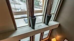 A room with a view of a mountain landscape through a large window. There is a small seating area with a lamp and a rug.