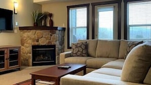 A living room with a stone fireplace, a beige sofa, a coffee table, and a flat-screen TV mounted on the wall.