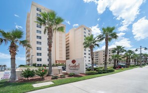 A hotel with a sign indicating private residential condominiums, a parking area, and a well-maintained lawn with palm trees. - Panoramic Views! Luxury 10th Floor Condo at Sanibel Condominium #1001 (Daytona Beach Shores)