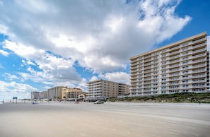 A beachfront hotel with multiple floors, balconies, and a sandy beach in the foreground.