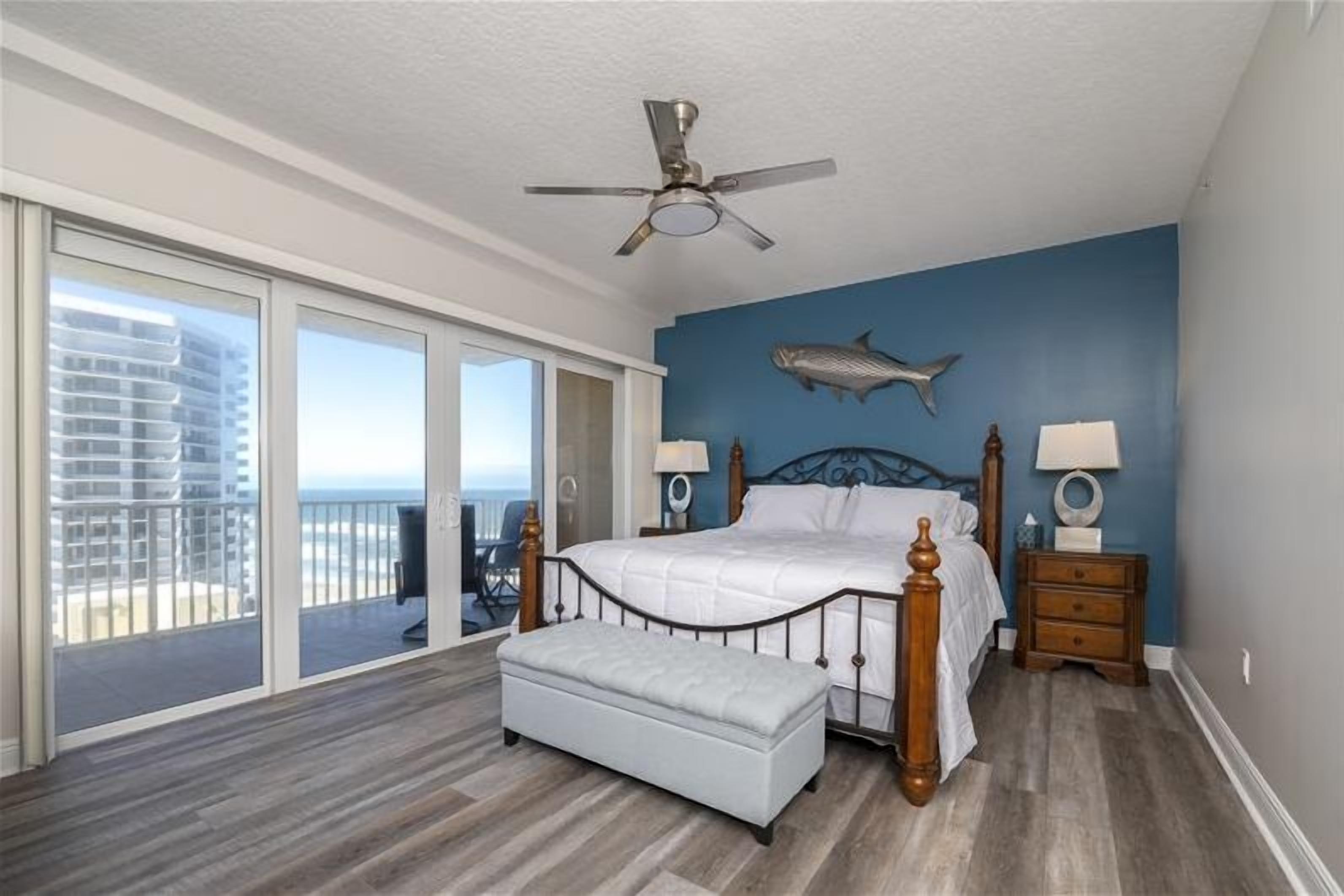 A bedroom with a large bed, a ceiling fan, a fish wall decoration, and a view of the ocean through a sliding door.
