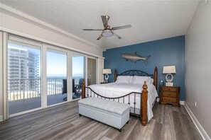 A bedroom with a large bed, a ceiling fan, a fish wall decoration, and a view of the ocean through a sliding door.