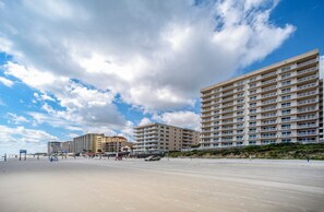 Ein Strandhotel mit mehreren Etagen, ein Sandstrand im Vordergrund und ein klarer Himmel mit Wolken.