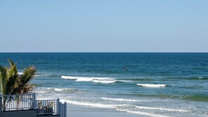A beach view with waves, a palm tree, and a fence. - Gorgeous 3/3 Direct Oceanfront First Floor at the Sanibel Condominium Unit #104 (Daytona Beach Shores)