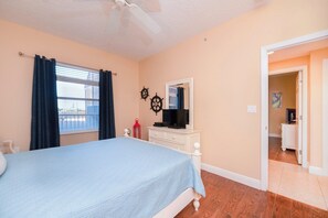 Une chambre à coucher comprenant un lit, une commode, un ventilateur de plafond, une fenêtre avec des rideaux et une porte donnant sur une autre pièce.