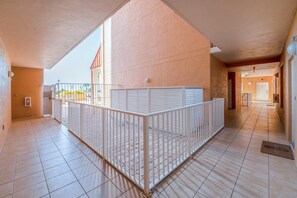 Un pasillo con barandilla de metal blanco, suelo de baldosas y un edificio con fachada color beige.