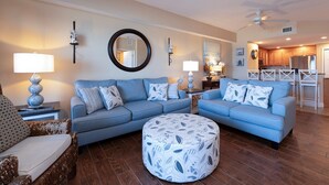 A living room with a blue sofa set, a round ottoman, and a dining area with white chairs. - Gorgeous 3/3 Direct Oceanfront First Floor at the Sanibel Condominium Unit #104 (Daytona Beach Shores)