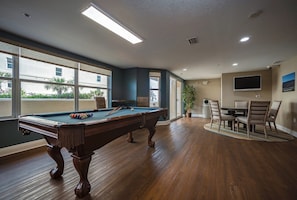 Una mesa de billar en una habitación con ventanas amplias, un comedor con sillas y un televisor en la pared.