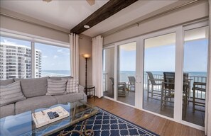 A spacious living room with a large window overlooking the ocean, a balcony with outdoor seating, and a modern interior design.
